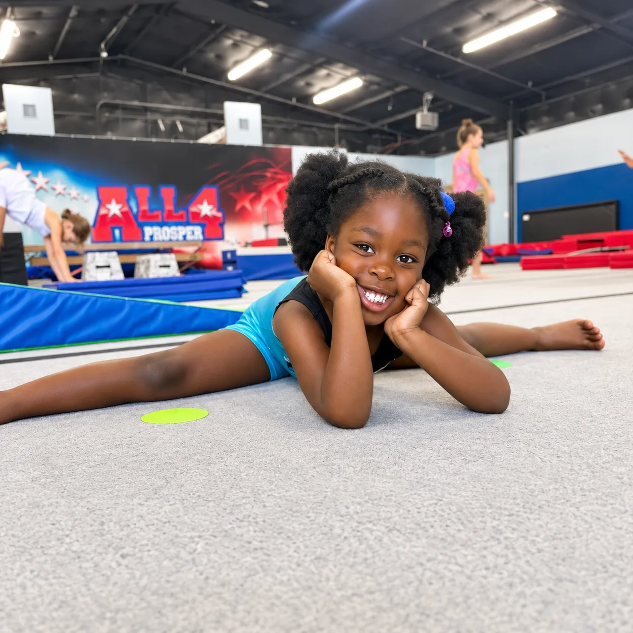 A smiling young gymnast holding a straddle stretch on the All 4 Prosper gym floor.